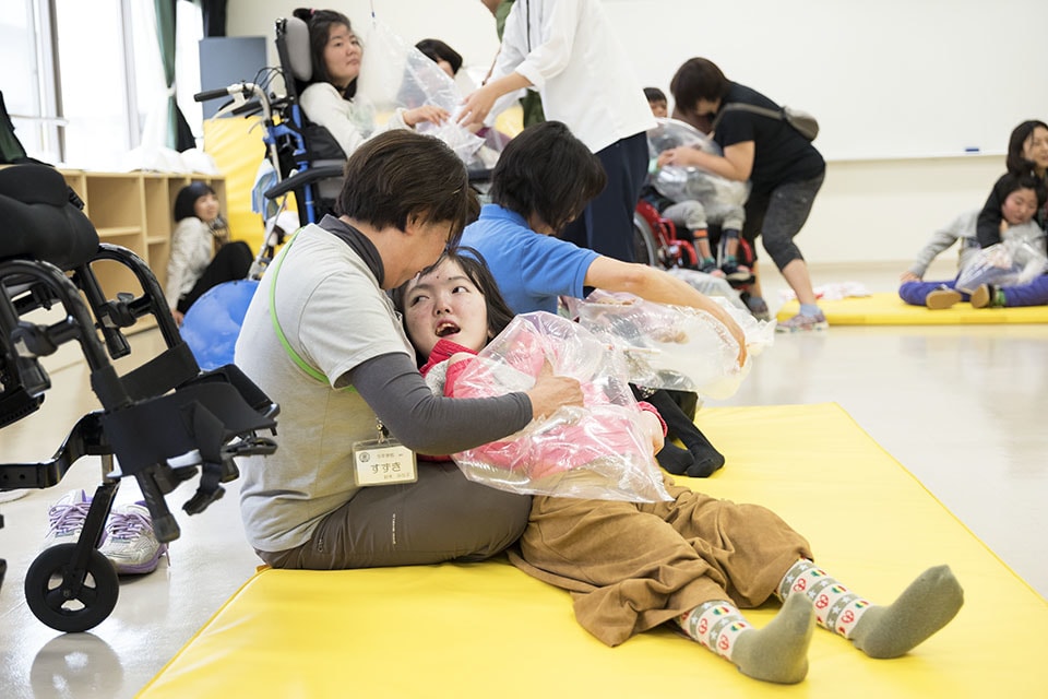 車椅子から降りて床に横たわりながら、風船のように膨らませたビニール袋を先生と一緒に鳴らす子ども