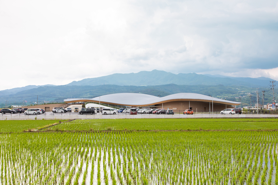 【Photograph】隣接する田園風景から臨む外観。背後の蔵王連峰に呼応した形状であることがわかる