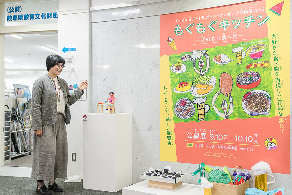 【写真】二村元子さんがギャラリーで開催中の企画展もぐもぐキッチンのポスターを紹介している