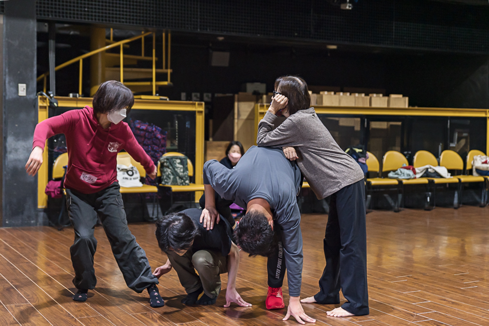 【写真】ダンスレッスンの一幕。ポーズをとる人、しゃがんでいる人、前かがみの人、その上にひじを突いて立っている人。奥には休んでいる人も。