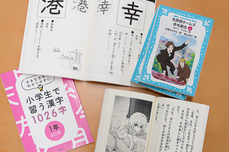 5冊の大きな文字の本。左下：大きな文字で「小学生で習う漢字1026字」と書名が書かれているピンクの表紙の本。右上：「名探偵ホームズ赤毛組合」という書名の青い表紙の本。左上：開いた本に大きく、幸（さいわい）、港（みなと）の漢字が書かれている。右下：開いた本にはふりがなのついた大きな文字の文章と挿絵がある。