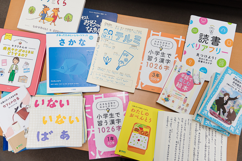 【写真】約15冊の本がテーブルの上に並べられている。さかなというタイトルの点字の絵本や、小学生で習う漢字の本、いないいないばあと描かれた触れる絵本、お弁当が表紙のわたしのおべんとうという本など。
