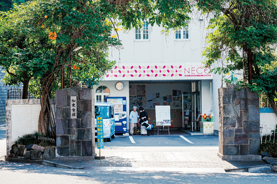 【写真】愛媛県・松山の〈和光会館〉外観。石造りの門の両脇に木が植えられ、地面に木漏れ日が映っている。門の先には駐車場と、同館に併設された、NPO法人シアターネットワークえひめが運営する〈シアターねこ〉の玄関口がある。